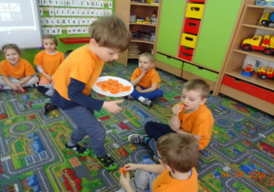 Dzieci siedzą na dywanie, chłopiec częstuje je marchewką.